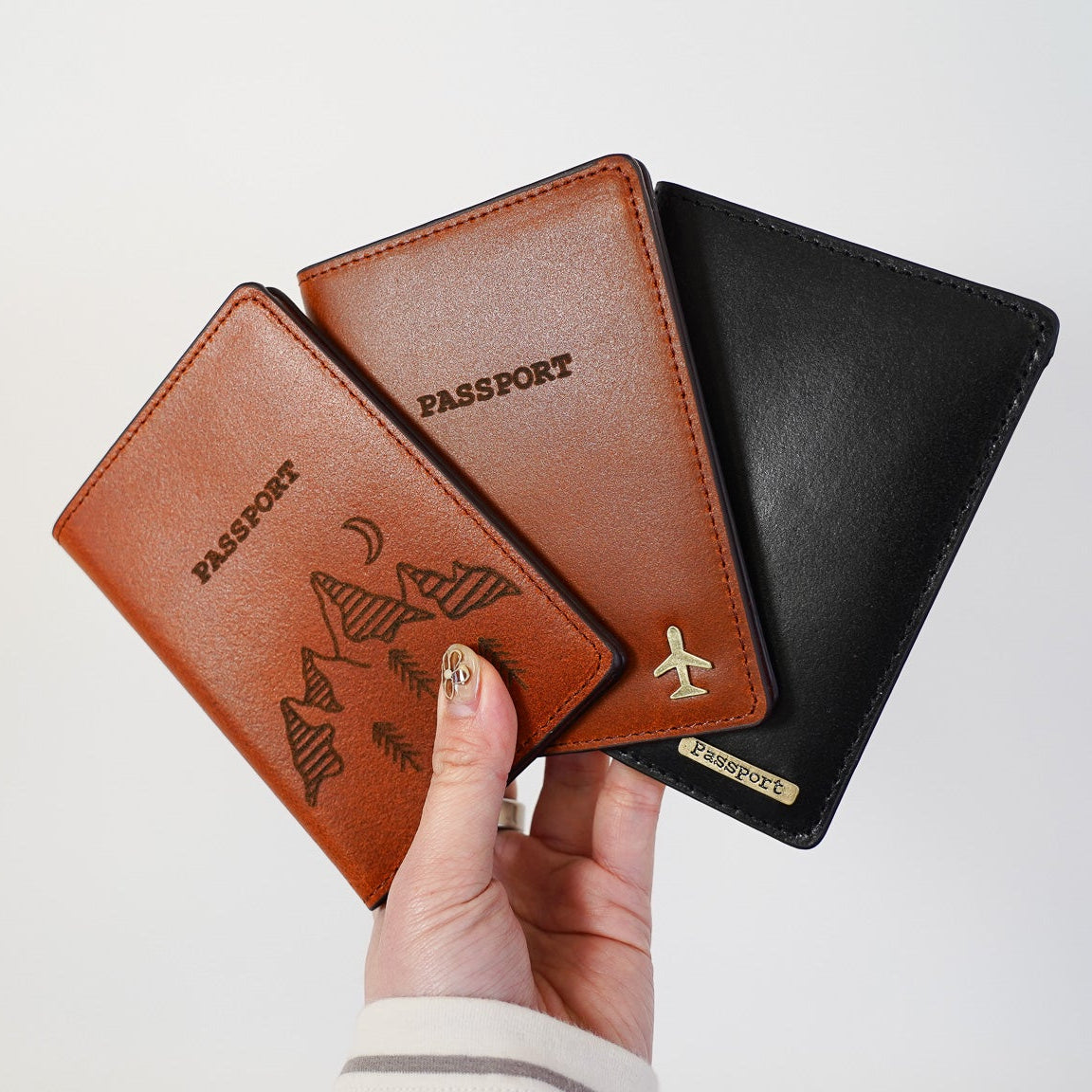 Three leather passport holders in brown, black, and tan held by a hand against a white background.