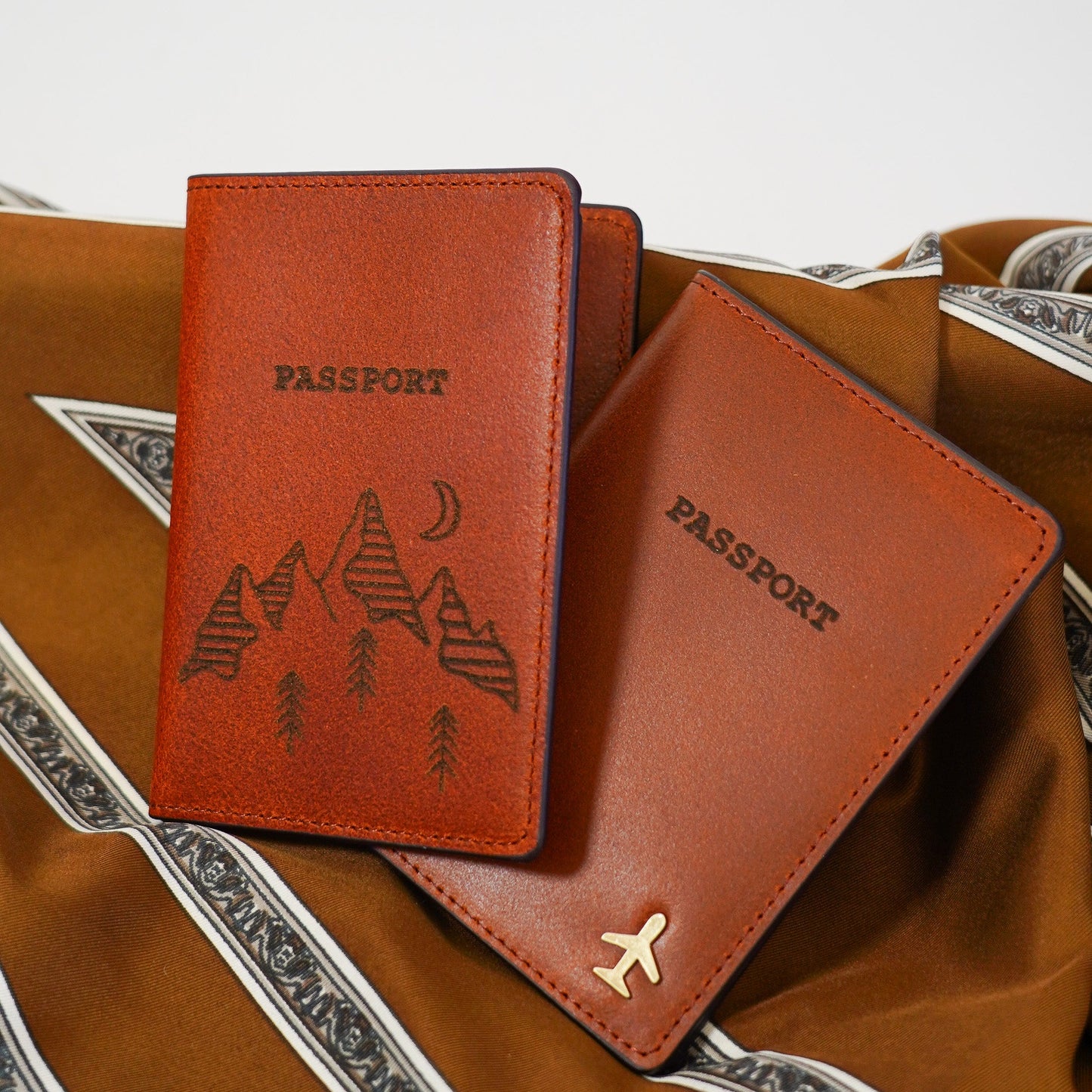 Two brown leather passport holders with mountain and airplane designs on a textured surface.