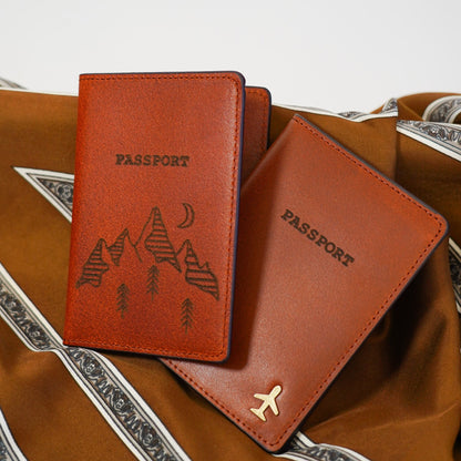 Two brown leather passport holders with mountain and airplane designs on a textured surface.